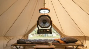 A photorealistic 4K image showing the Explore battery powered fan hanging from the ceiling of a bell tent with its integrated LED light on, casting soft illumination on Vango and Outwell sleeping gear.