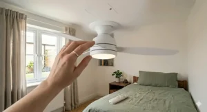 A person's hand holding a sleek white remote control to operate a modern bedroom ceiling fan and light.