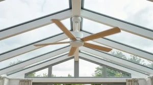 A classic 5-blade wooden ceiling fan with integrated LED light installed on the central ridge of a Victorian-style conservatory roof.