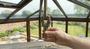 A hand holding a remote control unit to adjust the speed and dimmable light of a ceiling fan in a sunlit home conservatory.