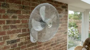 A hand holding a remote control to adjust a smart oscillating fan inside a bright, south-facing residential conservatory.