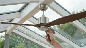 An oscillating wall-mounted fan fixed to the brick dwarf wall of a conservatory, directing airflow toward a dining table.