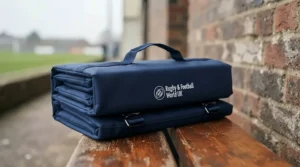 Compact foldable portable stadium cushion with an integrated carry strap resting on a wooden bench at a local UK sports ground.